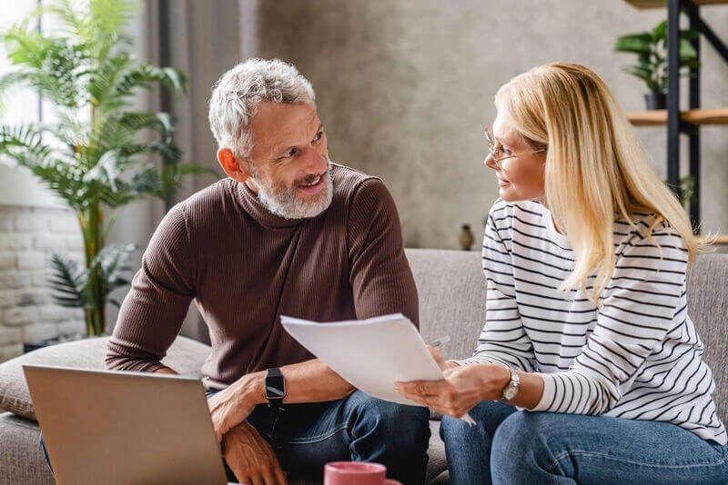 Couple souriant d'âge moyen avec un ordinateur portable étudiant des documents tout en travaillant sur le canapé à la maison
